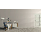 A minimalist room scene with a gray wall, light wood floor, a blue upholstered armchair with a draped throw, a small desk with a lamp and books, and a white door on the right.