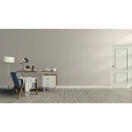 A minimalist room scene with a gray wall, light wood floor, a blue upholstered armchair with a draped throw, a small desk with a lamp and books, and a white door on the right.