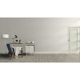 A minimalist interior room with a light gray wall, gray wood floor, a white door on the right, and a furnished study area on the left featuring a desk, lamp, chair, and small cabinet.