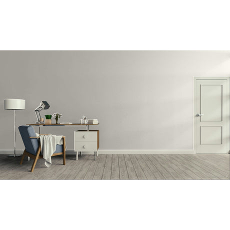 A minimalist interior room with a light gray wall, gray wood floor, a white door on the right, and a furnished study area on the left featuring a desk, lamp, chair, and small cabinet.