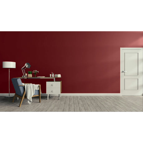 A modern room scene with a deep red wall, light gray wood floor, a white door on the right, and a home office setup on the left featuring a blue armchair, desk, lamp, and floor lamp. Prestige Hidden Notebook Paint