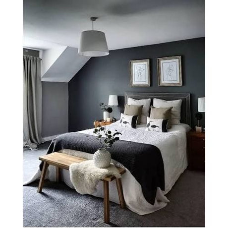 A styled bedroom interior featuring a bed dressed in white and black bedding, set against a dark gray wall with two framed prints. A wooden bench with a white faux-fur throw sits at the foot of the bed, and a gray rug covers the floor.