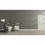 A minimalist room scene with a gray wall, light wood floor, a white door on the right, and a furnished study area on the left featuring a desk, lamp, chair, and draped throw blanket.
