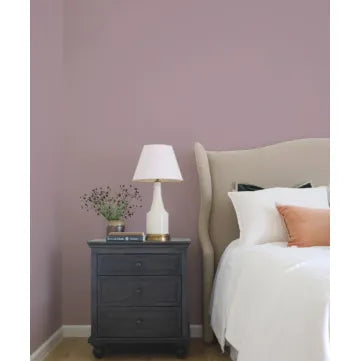 A styled bedroom corner featuring a dusty mauve-purple wall, a dark charcoal nightstand with a white table lamp and small vase of flowers, and part of a bed with white linens and a peach accent pillow.