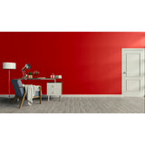 A modern room with a bold red wall, light gray wood flooring, a white door on the right, and a home office setup on the left featuring a desk, lamp, chair, and throw blanket.
