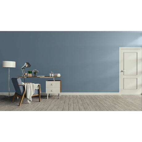 A minimalist room scene with a muted blue-gray wall, light wood floor, a white door on the right, and a desk setup on the left including a chair, lamp, and small cabinet.