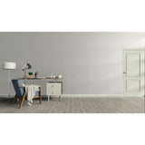 A minimalist interior room scene with a light gray wall, gray wood floor, a white door on the right, and a home office setup on the left featuring a desk, lamp, chair, and throw blanket.