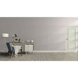 A minimalist room scene with a light gray wall, gray wood floor, a white door on the right, and a home office setup on the left featuring a desk, lamp, chair, and small cabinet.