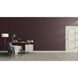A modern interior room with a dark plum-brown accent wall, light wood flooring, a white door on the right, and a home office setup on the left featuring a desk, lamp, chair, and throw blanket.