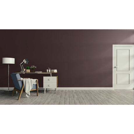 A modern interior room with a dark plum-brown accent wall, light wood flooring, a white door on the right, and a home office setup on the left featuring a desk, lamp, chair, and throw blanket.