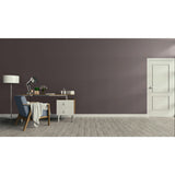 A modern interior room scene with a dark mauve-grey wall, light wood floor, white door on the right, and a home office setup on the left featuring a desk, lamp, chair, and throw blanket. Prestige Mustang Paint