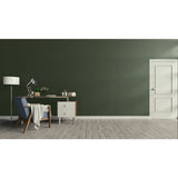 A modern room scene with a dark olive green wall, light wood plank flooring, a white door on the right, and a home office setup on the left featuring a desk, lamp, chair, and throw blanket.