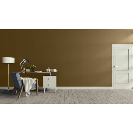 A modern interior room scene with a large warm brown wall, light wood floor, white door on the right, and a furnished home office area on the left featuring a desk, lamp, chair, and small cabinet.