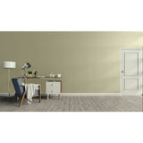 A minimalist room scene with a beige wall, gray wood floor, white door, a blue armchair with a draped throw, a desk with a lamp and small plants, and a floor lamp on the left.