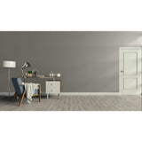 A minimalist room scene with a gray wall, light wood floor, a white door on the right, and a furnished study area on the left featuring a desk, lamp, chair, and throw blanket.
