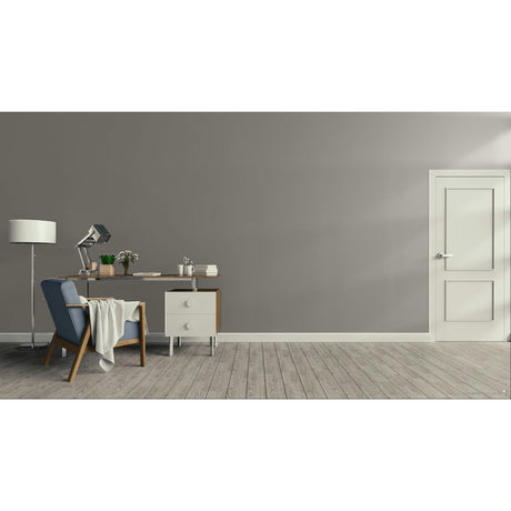 A minimalist room scene with a gray wall, light wood floor, a white door on the right, and a furnished study area on the left featuring a desk, lamp, chair, and throw blanket.