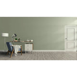 A minimalist room scene with a sage green wall, light wood floor, a white door on the right, and a home office setup on the left featuring a desk, lamp, chair, and throw blanket.