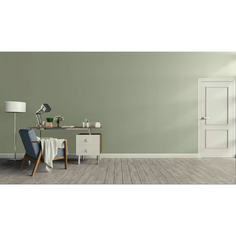 A minimalist room scene with a sage green wall, light wood floor, a white door on the right, and a home office setup on the left featuring a desk, lamp, chair, and throw blanket.