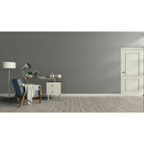 A minimalist room scene with a gray wall, light wood floor, white door, desk, chair, lamp, and small cabinet.