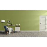 A styled interior room with a sage green wall, gray wood floor, a white door on the right, and a home office setup on the left featuring a blue armchair, desk, lamp, and small cabinet.