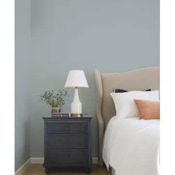 A styled bedroom corner featuring a dark charcoal gray three-drawer nightstand next to a bed with white linens, a beige wingback headboard, and a white table lamp with a beige shade.