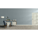 A minimalist room scene with a muted blue-gray wall, light wood floor, a white door on the right, and a furnished study area on the left with a blue armchair, desk, lamp, and small cabinet.