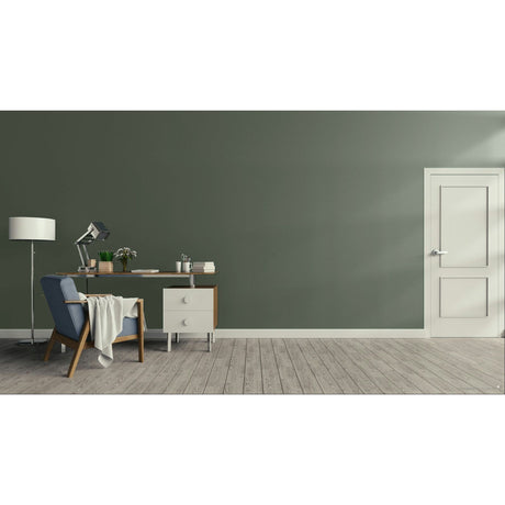 A minimalist room scene with a dark green wall, light wood floor, a white door on the right, and a home office setup on the left featuring a desk, lamp, chair, and throw blanket.