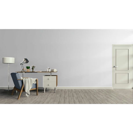 A minimalist room scene with a light gray wall, wood floor, a blue upholstered armchair with a draped throw, a wooden desk with a lamp and small accessories, and a cream-colored door on the right.