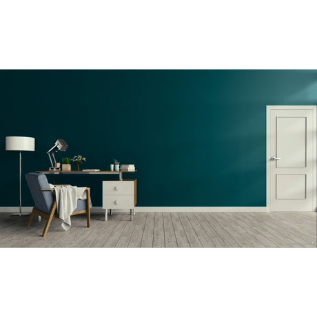 A modern interior scene with a deep teal wall, light wood floor, a white door on the right, and a home office setup on the left featuring a desk, lamp, chair, and draped throw blanket.