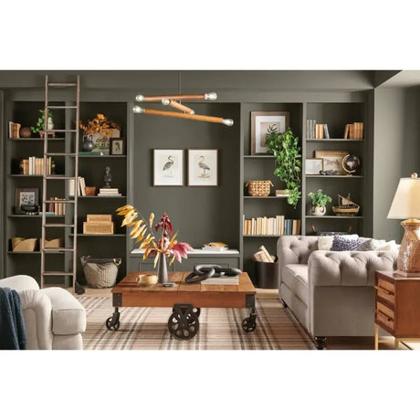 A styled interior room scene with built-in dark green shelves, books, plants, framed art, a wooden ladder shelf, a tufted gray sofa, a coffee table, and warm ambient lighting.