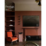 An interior room scene showing a terracotta-colored wall with built-in shelving, a black wall-mounted artwork, a modern orange armchair, and a glossy black table. The Prestige Fine Paint logo appears in the upper left corner.
