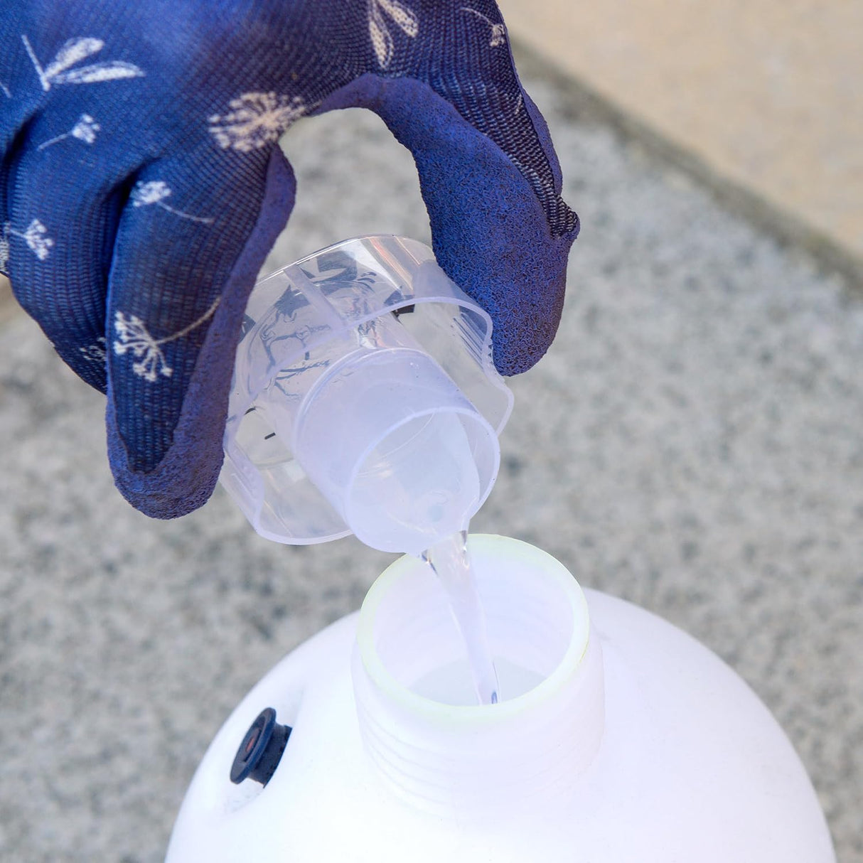 Hand wearing a blue glove pouring liquid from a small container into a larger white container on a concrete surface.