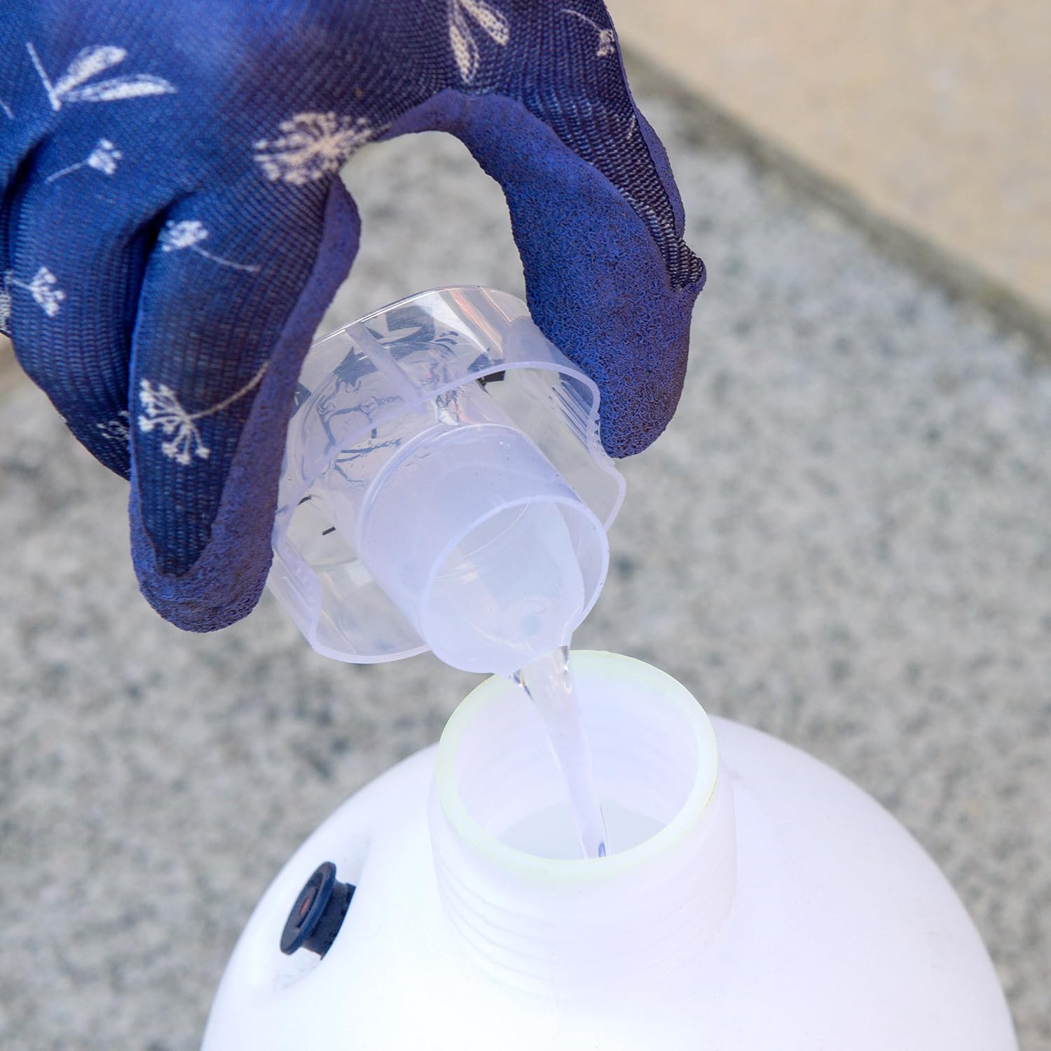 Hand wearing a blue glove pouring liquid from a small container into a larger white container on a concrete surface.