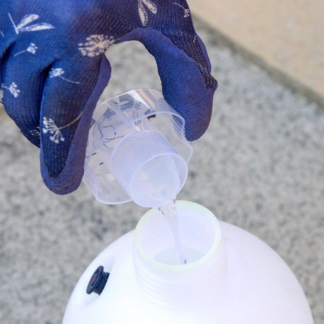 Hand wearing a blue glove pouring liquid from a small container into a larger white container on a concrete surface.