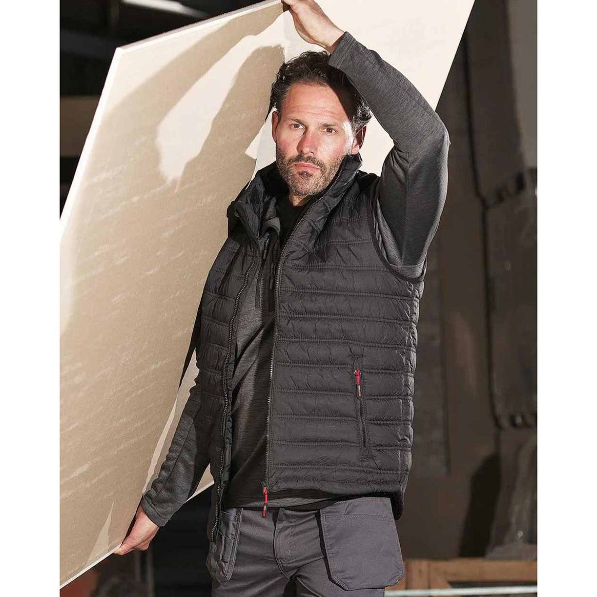 A man wearing a dark grey quilted puffer vest over a black shirt and gray cargo pants, holding a large beige board overhead in a workshop setting. Tuffstuff Elite Bodywarmer