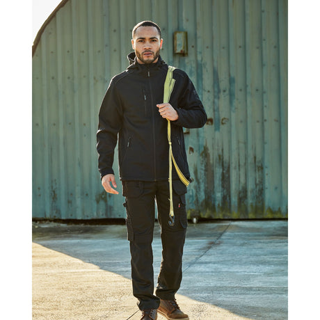 A man wearing a black hooded jacket with a bright yellow-green strap detail, black cargo pants, and brown boots, standing in front of a weathered teal metal wall. TuffStuff Hale Jacket