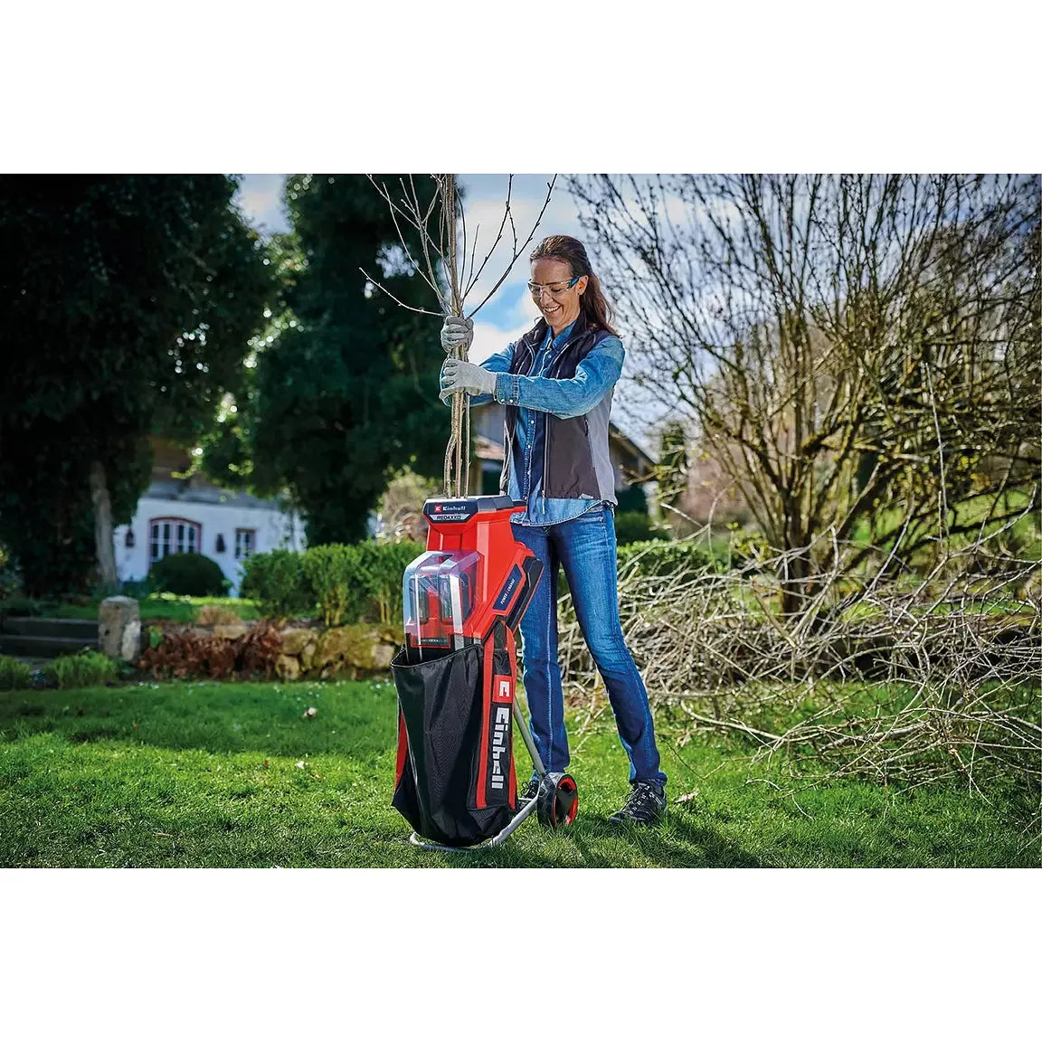 A woman stands in a grassy backyard holding a red and black leaf vacuum cleaner with a clear dust bin. The vacuum has the brand name 'BULLSKT' visible on the side.