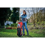 A woman stands in a grassy backyard holding a red and black leaf vacuum cleaner with a clear dust bin. The vacuum has the brand name 'BULLSKT' visible on the side.