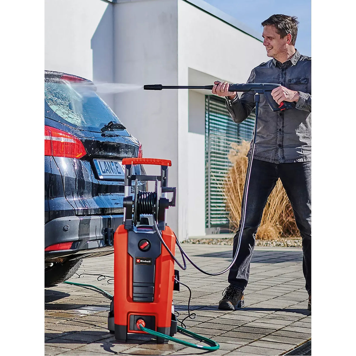 A man is washing a black SUV outdoors using a red and black K&N brand high-pressure water cleaner with a hose on a paved driveway. Einhell Power Tool Classic Pressure Washer 1900W 140 Bar