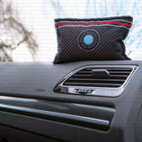 A car air freshener pillow with a carbon fiber pattern, featuring a circular blue and white logo that reads 'BENZIN AIR INTERNATIONAL', resting on a car dashboard next to an air vent.