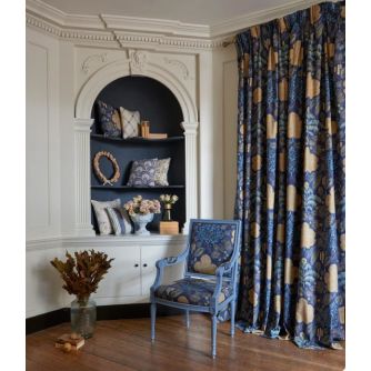 Prestige Ando
A styled interior room showing a pair of floor-length curtains in a blue and beige botanical pattern, next to a blue upholstered armchair and a built-in white shelf with decorative pillows.