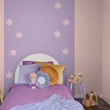 A styled children's bedroom corner with a lavender-purple wall decorated with white sun decals, a white bed frame, purple bedding, and decorative pillows including a flower-shaped pillow and a doll.