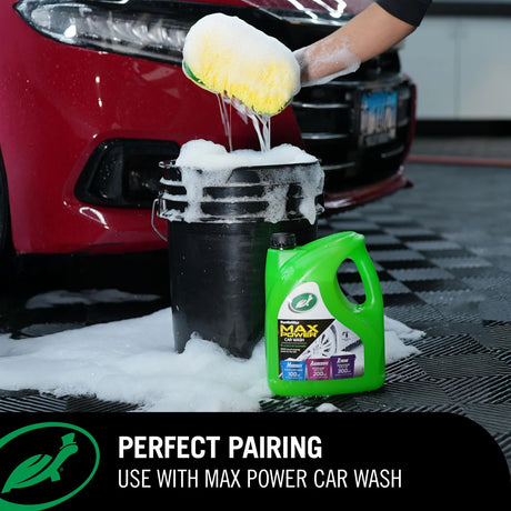 A green bottle of car wash liquid stands next to a black bucket filled with soapy water, with a person using a yellow sponge to scrub a red car in a garage setting. Text at the bottom reads 'PERFECT PAIRING USE WITH MAX POWER CAR WASH'. Turtle Wax M.A.X Power™ 3-in-1 Wash Pad