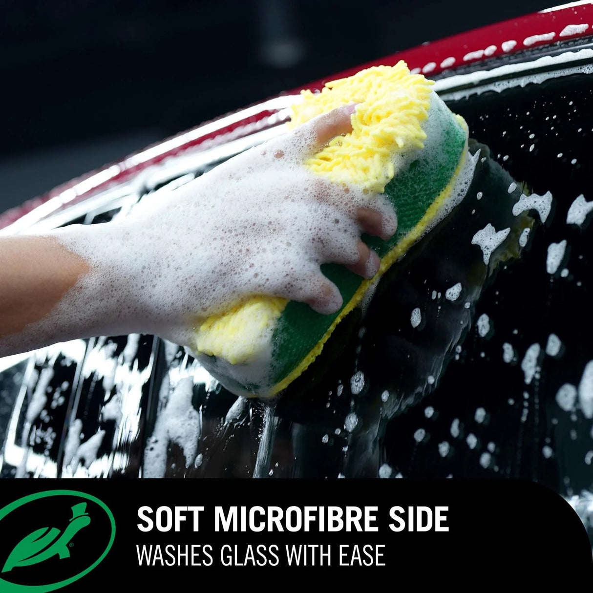 A hand wearing a white foam glove holds a green and yellow microfiber car wash sponge against a soapy black car window. A black banner at the bottom features a green logo and the text 'SOFT MICROFIBRE SIDE WASHES GLASS WITH EASE'.