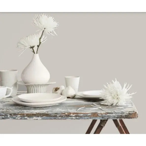 Prestige Hepburn Pale
A styled tabletop arrangement with matte white ceramic dishes, cups, a vase with white flowers, garlic bulbs, and a large white flower on a distressed rustic table against a plain beige wall.
