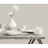 Prestige Hepburn Pale
A styled tabletop arrangement with matte white ceramic dishes, cups, a vase with white flowers, garlic bulbs, and a large white flower on a distressed rustic table against a plain beige wall.