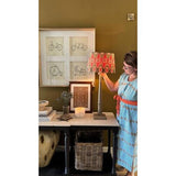b A woman in a blue and white patterned dress with a red waist sash stands next to a table displaying a lit table lamp with a red, orange, and black geometric patterned shade.