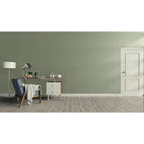 A minimalist room scene with a sage green wall, light wood floor, a white door on the right, and a home office setup on the left featuring a desk, lamp, chair, and throw blanket.