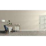 A minimalist interior room scene with a beige wall, gray wood floor, a white door on the right, and a home office setup on the left featuring a desk, lamp, chair, and throw blanket.