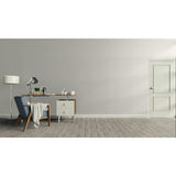 A minimalist room scene with a light gray wall, gray wood floor, a white door on the right, and a home office setup on the left featuring a blue armchair, desk, lamp, and side cabinet.